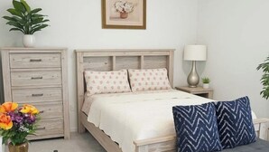 Quarto com cama de madeira, mesa de cabeceira com abajur, sofá cinza, pequena mesa de cabeceira com planta e um vaso de flores em um aparador.