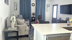 Uma sala de estar moderna com um sofá, um espaço de refeições com bancada de cozinha e uma televisão grande fixada na parede.
