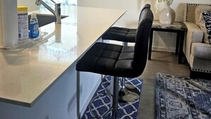Cozinha moderna com ilha central, pia e janela com cortinas azuis. Na área de estar, há um sofá e uma pequena mesa com um vaso de flores.