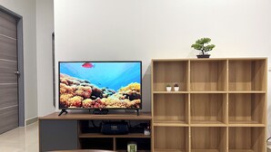 Una sala de estar con un televisor de pantalla plana sobre un soporte de madera, una mesa de centro con controles remotos, una estantería y una planta pequeña en la parte superior.