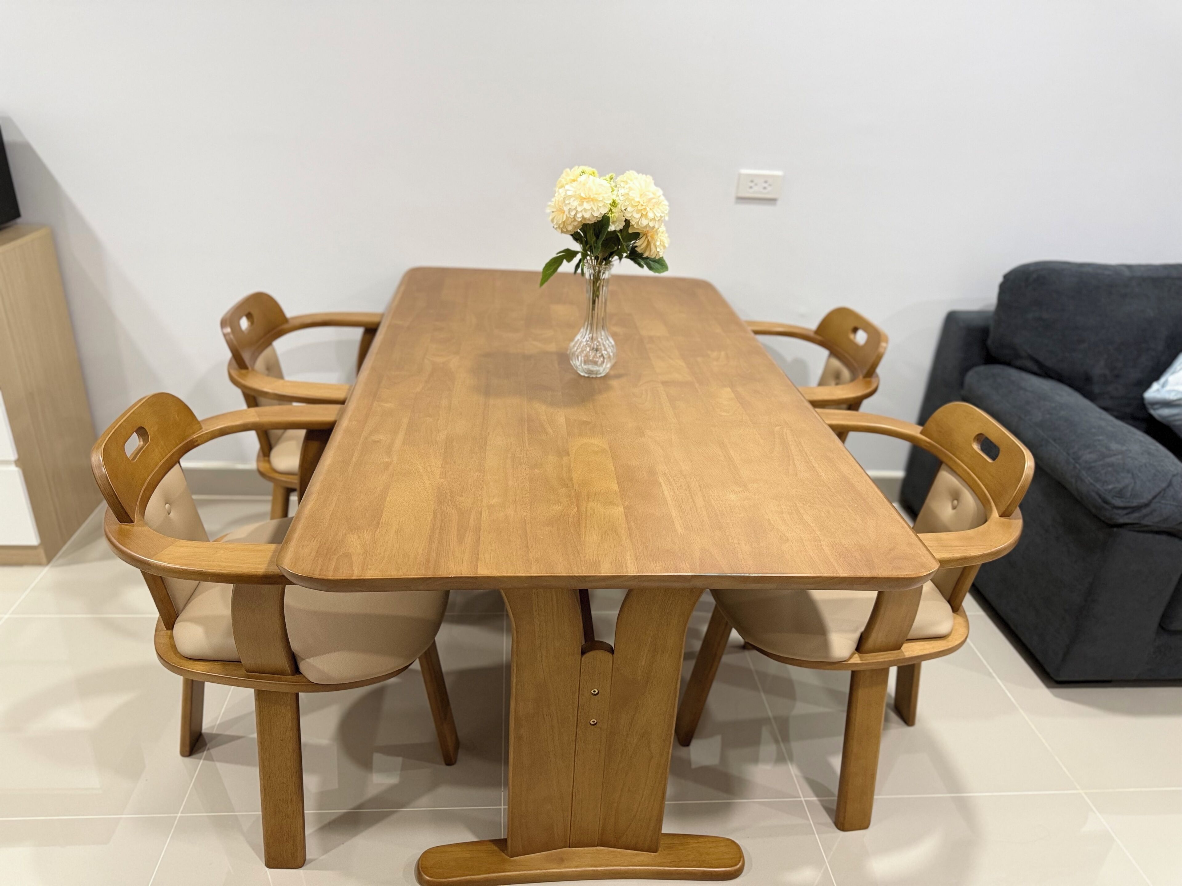 A wooden dining table with six chairs, a vase with flowers, and a grey sofa in the background.