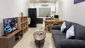 Una sala de estar moderna con un televisor de pantalla plana, estanterías de madera, un refrigerador negro, un sofá gris con cojines estampados, una mesa de centro de madera y un comedor con una mesa y sillas blancas.