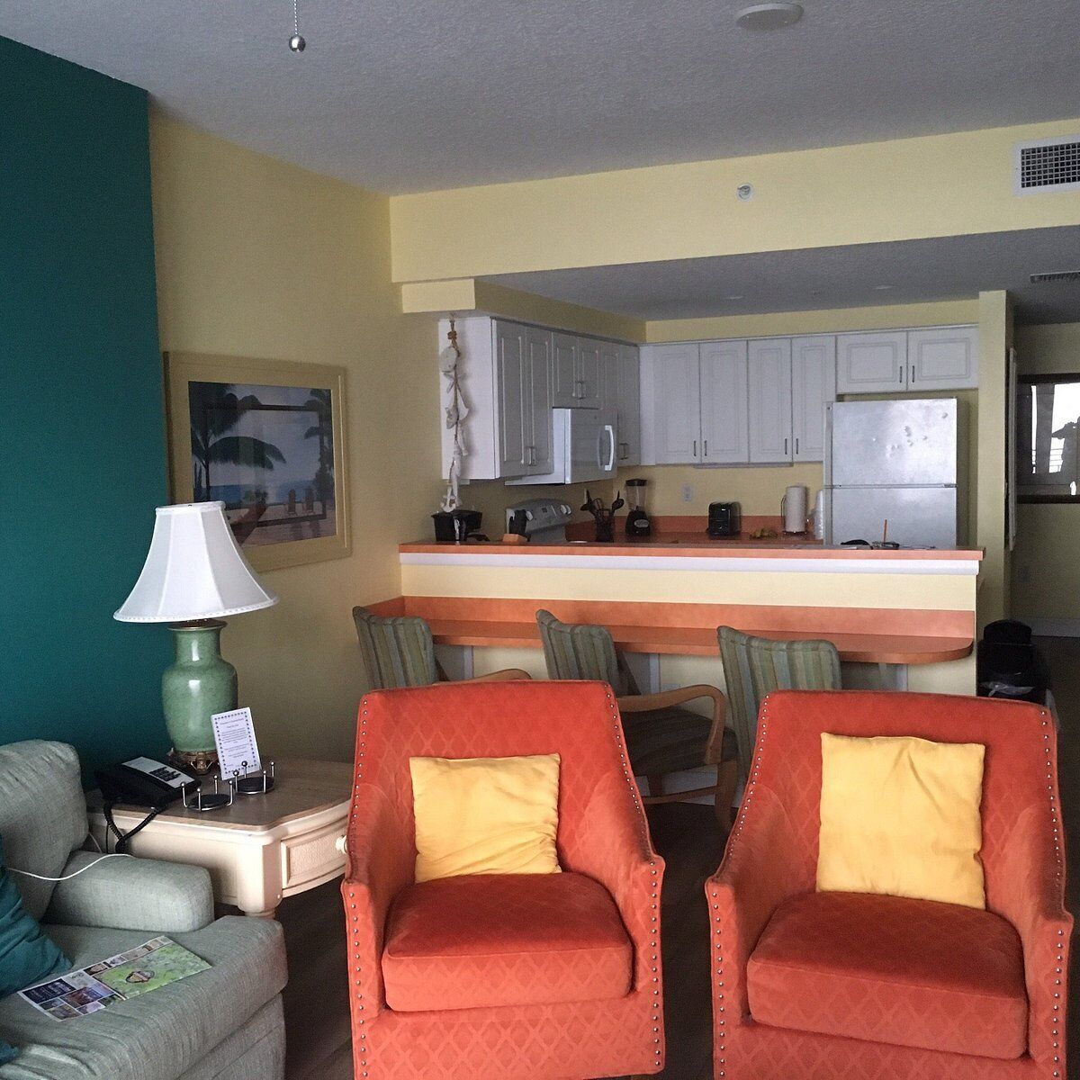 A compact living space with a kitchenette, two armchairs with yellow cushions, a small table with a lamp, and a framed picture on the wall.