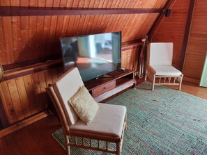 Ein gemütliches Zimmer mit Holzwänden und schrägverdeckter Decke, mit einem Fernseher auf einem Holzständer, zwei Korbstühlen und einem gewebten Teppich.