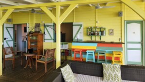 A room with a wooden floor, a sofa with cushions, a wooden cabinet, a bar area with stools, and a door with a green frame. - Villa, lodge 6-7 pers, exceptional view, comfort and nature from the center. (Le Diamant)