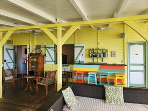 Ein Zimmer mit Holzboden, einer Couch mit Kissen, einem Holzkasten, einer Bar mit Hockern und einer Tür mit grünem Rahmen.