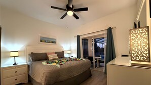 Un dormitorio con cama, mesitas de noche, ventilador de techo, un cuadro y vistas a través de un ventanal.