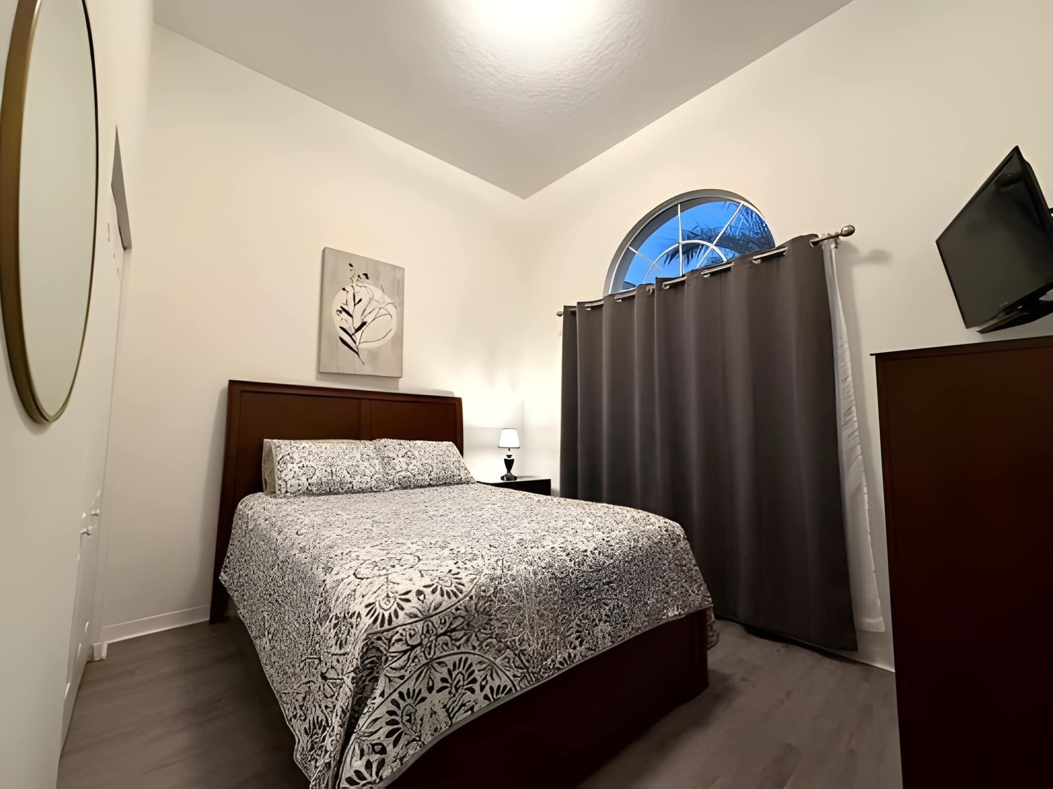 A bedroom with a bed, a nightstand, a wall-mounted TV, a window with curtains, and a wall art piece.