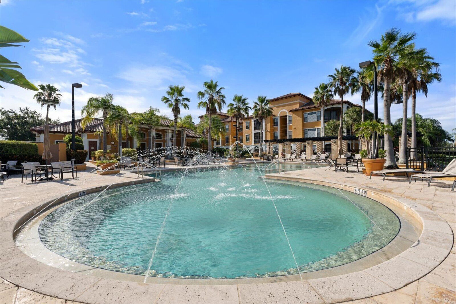 A circular pool with a water fountain, surrounded by lounge chairs and palm trees, with buildings in the background.