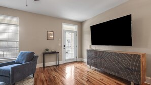 A living room with hardwood flooring, a blue armchair, a TV mounted on a patterned console, and a door with blinds.