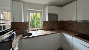 Une cuisine moderne avec des armoires blanches, un plan de travail en bois, un four encastré et une fenêtre donnant sur la forêt.
