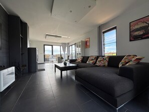 A modern living room with a dark sofa, a coffee table, and a view through large windows. - Enjoyable 3-bedroom house with Saun in Belgian Ardenne for your getaway (Fauvillers)