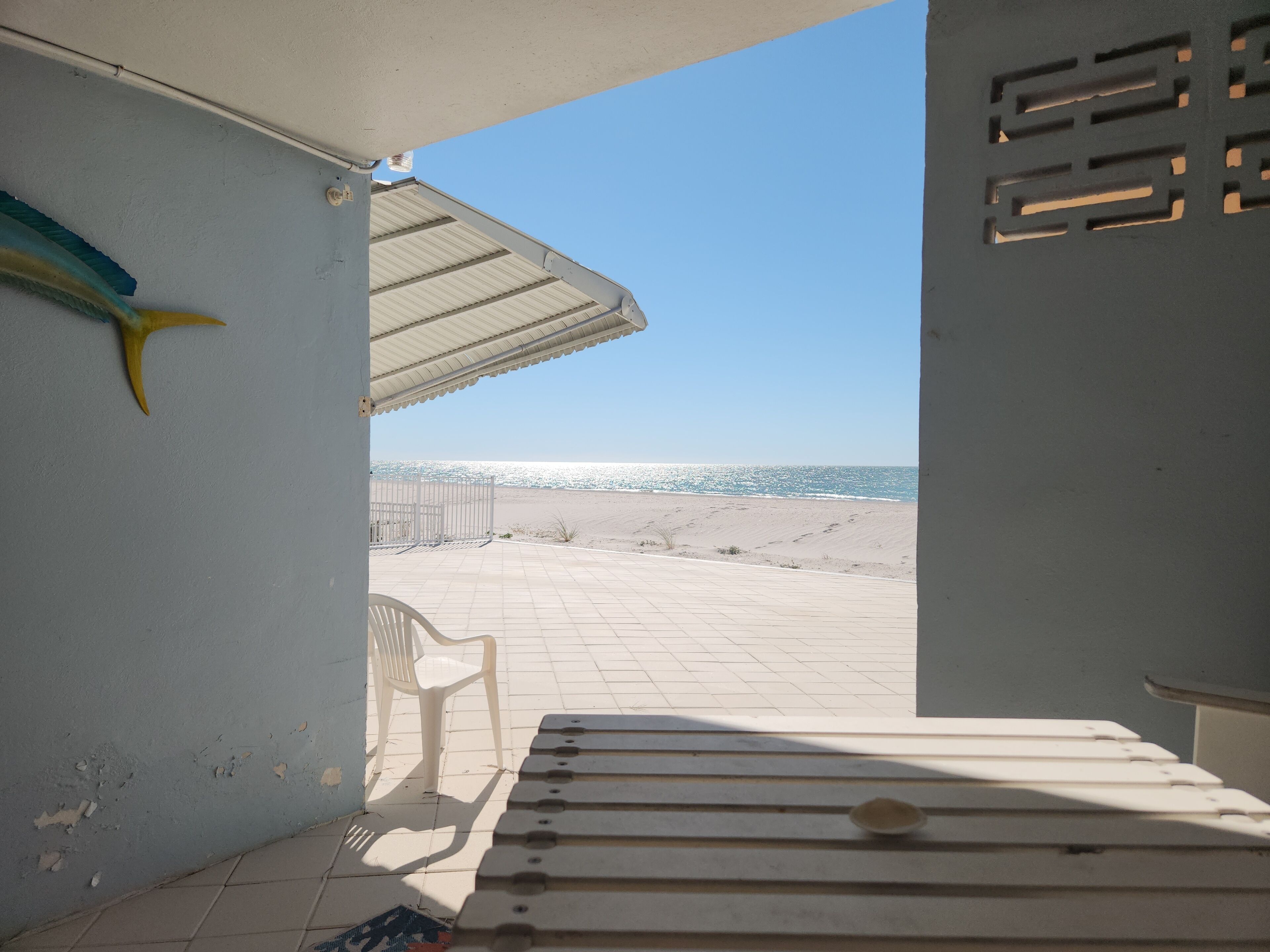 Vista de la playa desde un porche cubierto con un banco, una silla de plástico y un pez decorativo.