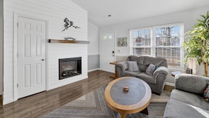 A living room with a fireplace, a ceiling fan, and a large window with blinds.
