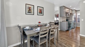 A modern dining area with a wooden table set for four, framed artwork on the wall, and a kitchen in the background.