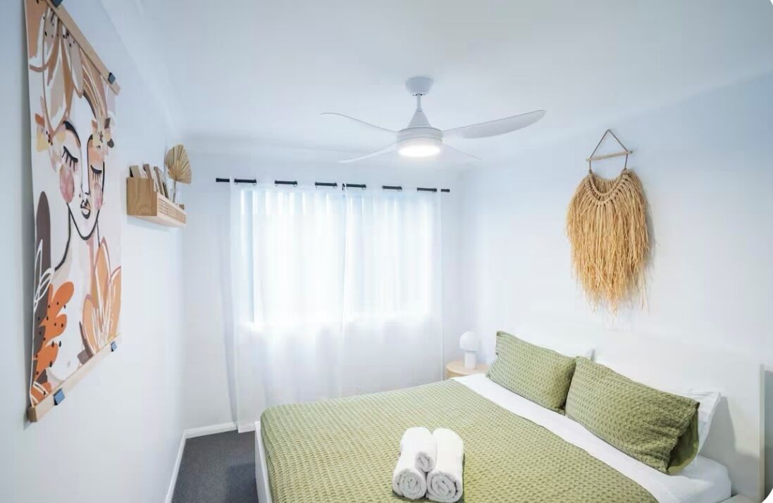 A hotel room with a bed, a ceiling fan, a window with curtains, and a wall-mounted macrame decoration.