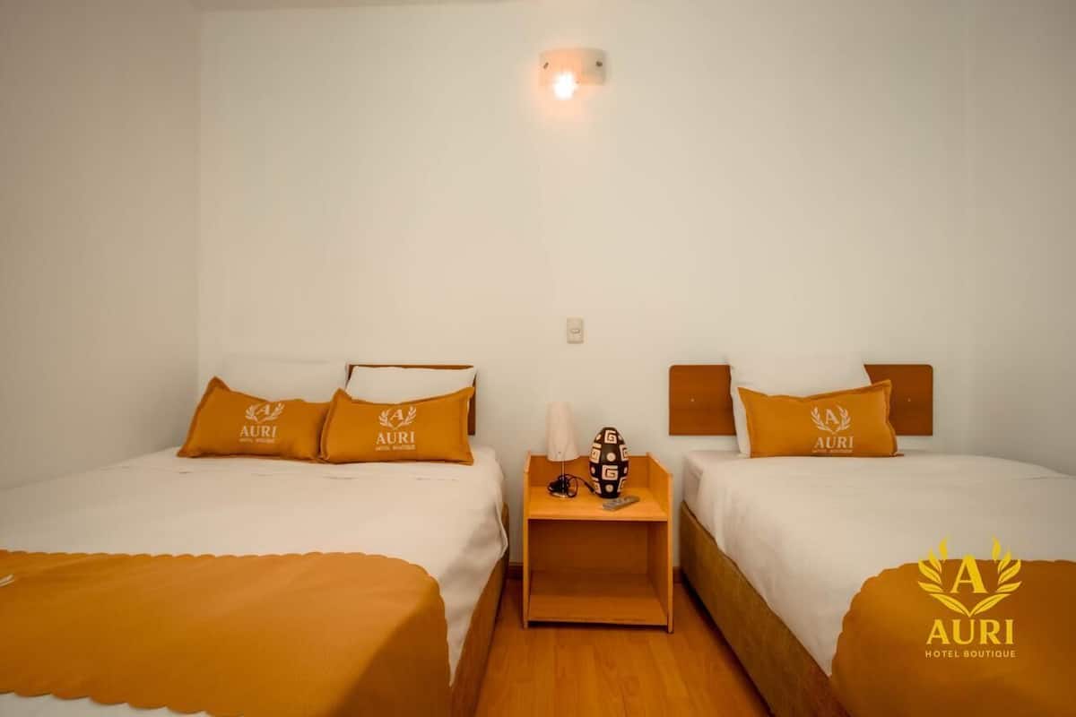 A hotel room with two beds, each with matching orange bedding and pillows, a nightstand with a lamp and a decorative item, and a wall-mounted light fixture.