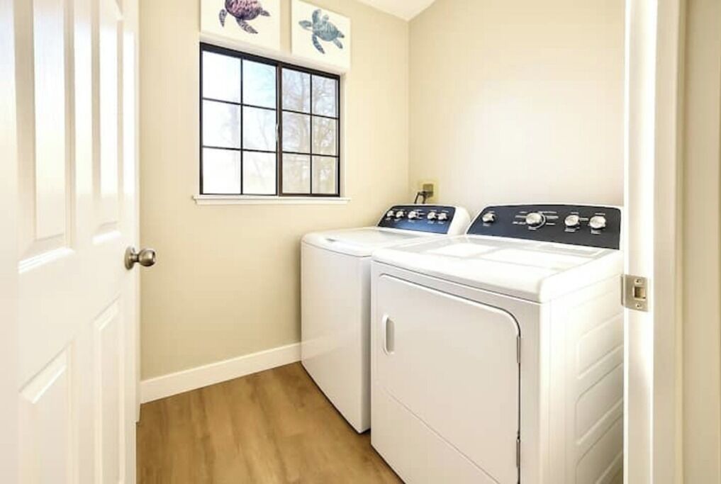 A laundry room with two white front-loading washers and dryers, a window with a view of trees, and a door on the left.