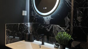 Un bany amb una paret de marbre negre, un mirall rodó amb llum, un lavabo blanc i una planta en test.