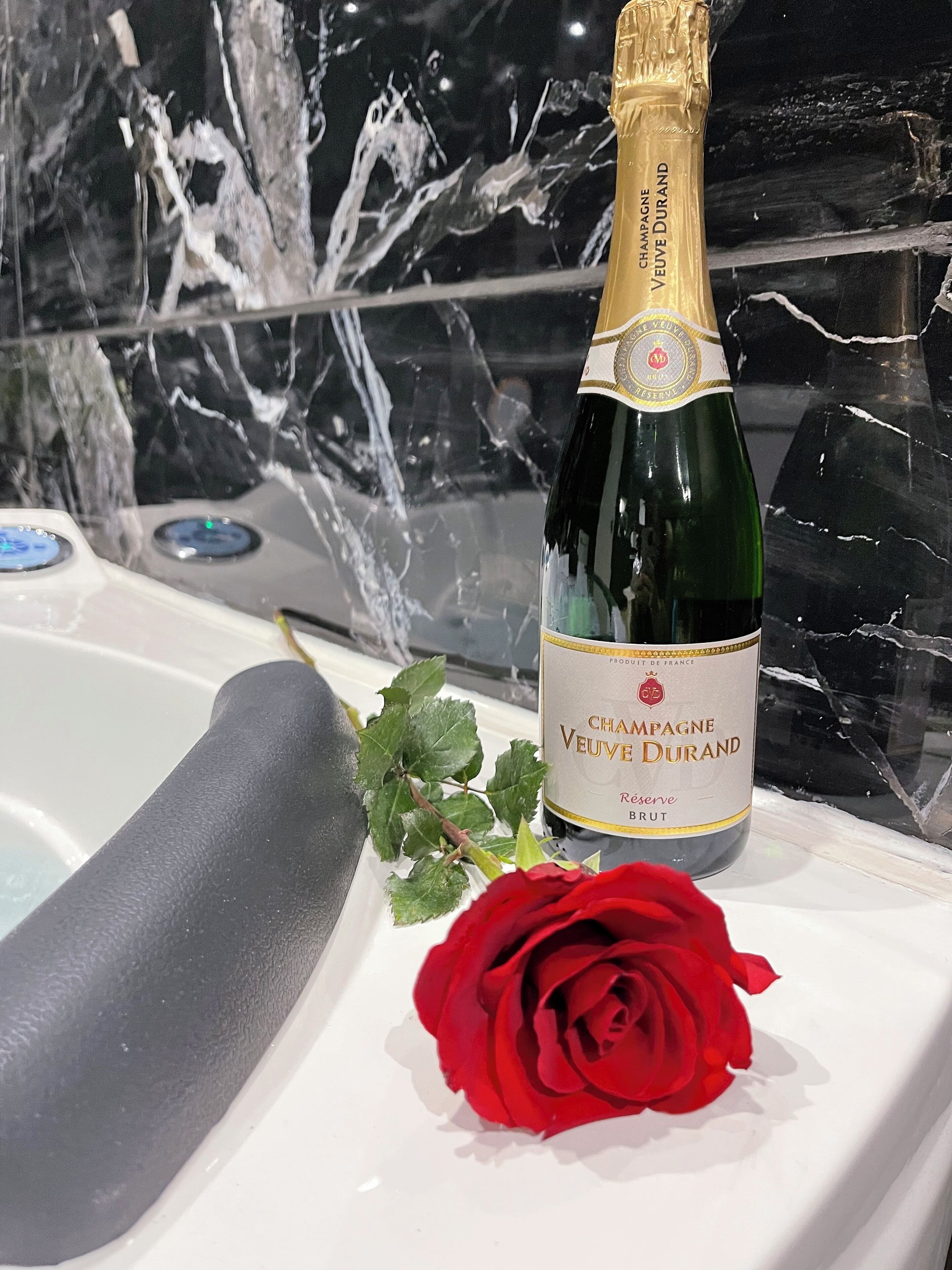 Valkoisella alustalla on pullo Champagne Veuve Durand Reserve Brut -kuohuviiniä, ja sen vieressä on punainen ruusu.