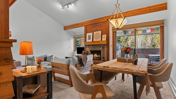 A cozy living room with a fireplace, a dining area with a wooden table and chairs, and a large window with a view of the outdoors. - RMR: Alpine Fir 722 Condo in The Aspens/Wilson, WY (Wilson)