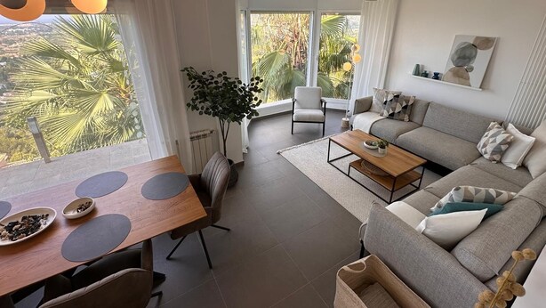 Ein modernes Wohnzimmer mit einer Couch, einem Esstisch und Stühlen. Ein großes Fenster bietet Blick auf Palmen und es gibt einen Deckenventilator.