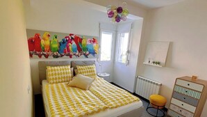 Ein Schlafzimmer mit Bett, Schminktisch, Stuhl, Heizkörper, Fenster und einem Bild bunter Vögel.