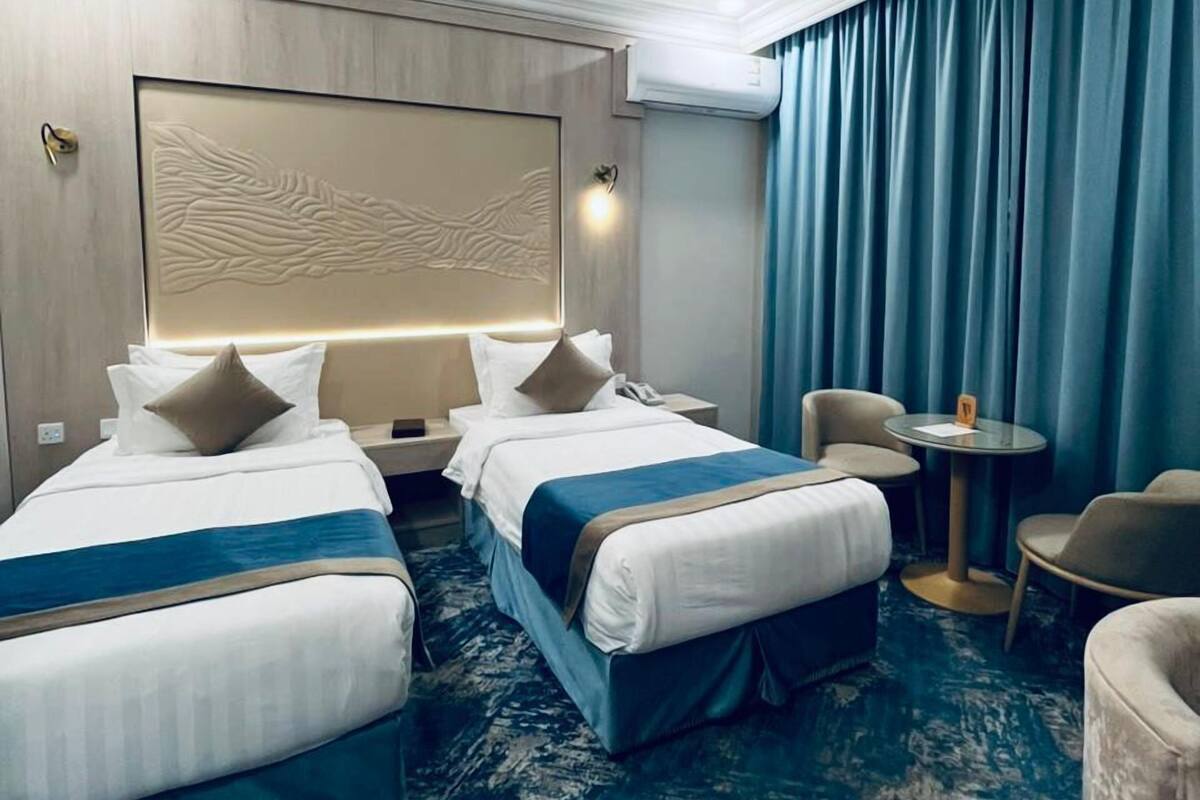 A hotel room with two beds, a small round table, a chair, and a large window with curtains.