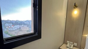 A hotel room with a large window, a bedside table with a phone, and a bed with white linens. - The High View (Al Baha)