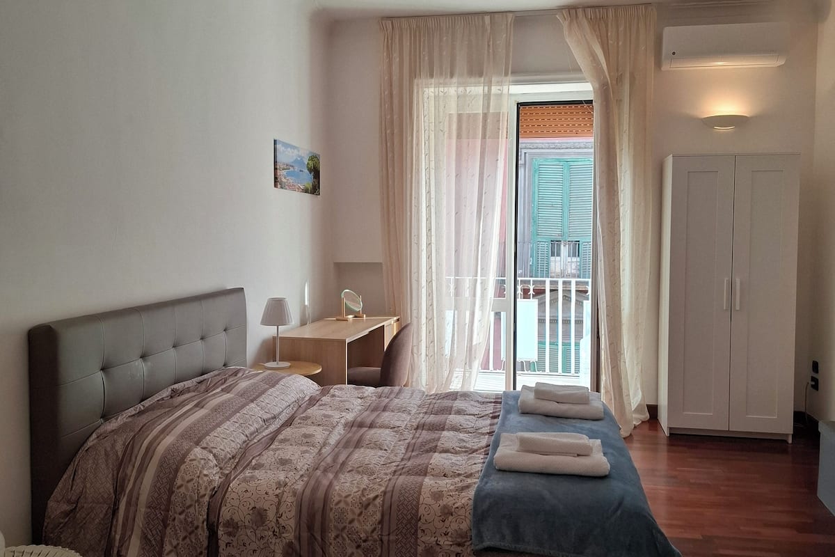 A bedroom with a bed, a desk, a chair, a wardrobe, and a view of the outside.