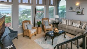 Un salon spacieux comprenant un canapé, des fauteuils, une table basse et un téléviseur. De grandes fenêtres offrent une vue sur le paysage.