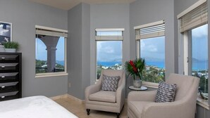 Une chambre à coucher comprenant un lit, deux fauteuils, une petite table et de grandes fenêtres donnant sur un beau paysage.
