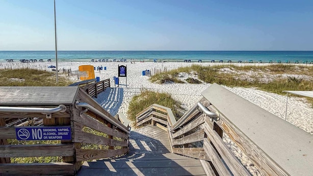 Ein hölzerner Steg führt zu einem Strand, mit einem Schild, das Rauchen, Dampfen, Alkohol, Drogen und Waffen verbietet.