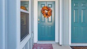 Uma porta azul-esverdeada com uma guirlanda decorativa, uma janela com persianas e um tapete de entrada vermelho.