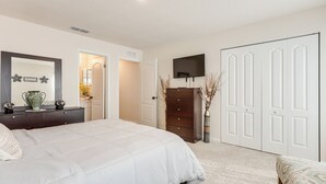 Quarto com cama grande, penteadeira, televisão montada acima de um guarda-roupa e porta que dá para outro cômodo.