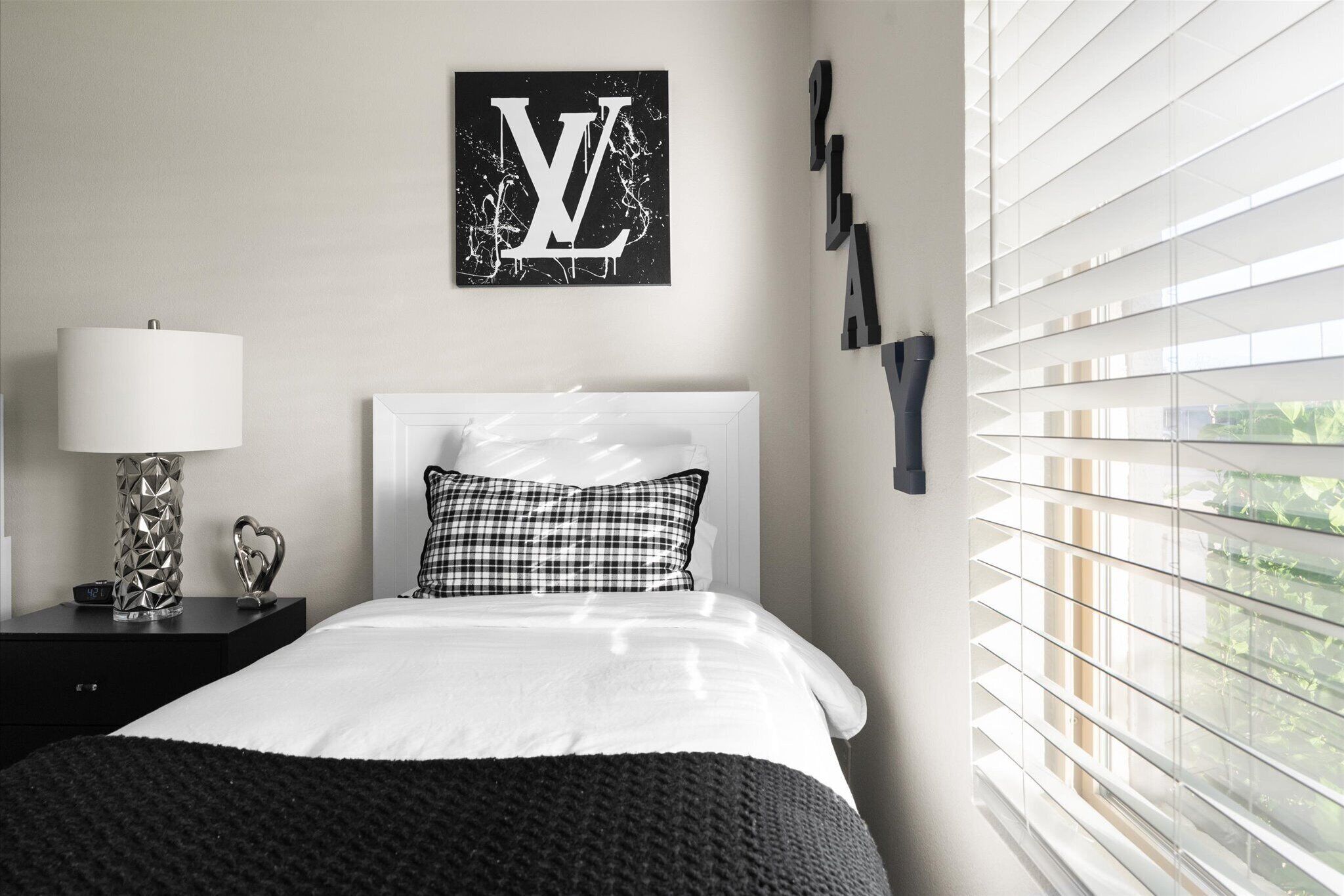 Une chambre à coucher avec un lit, une table de chevet avec une lampe, une œuvre d’art accrochée au mur et une fenêtre avec des stores.