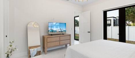 Quarto moderno com uma cama grande, uma TV sobre uma cômoda de madeira, e vista para o exterior por uma porta deslizante.