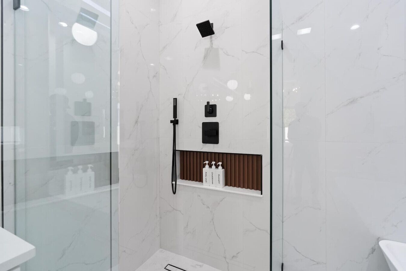 A modern bathroom with a glass shower enclosure, a built-in shelf with toiletries, and a marble wall.