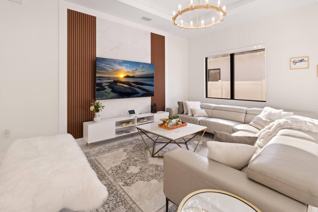 Una sala de estar moderna con un televisor de pantalla plana grande, un sofá color beige, un sillón blanco y una mesa de centro con objetos decorativos.