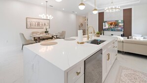 Una cocina moderna con una isla central, un comedor con mesa y sillas, y una sala de estar con sofá y televisor.