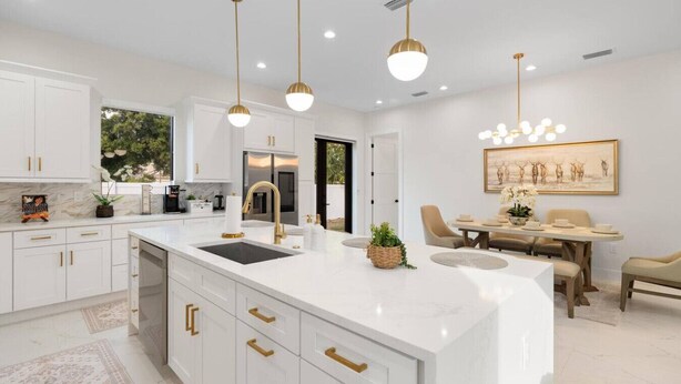 Una cocina moderna con armarios blancos, detalles dorados y una isla grande con fregadero. Hay un comedor con mesa y sillas, y un cuadro en la pared.