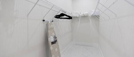 A small, white-walled room with a shower area, a towel rack, and a small shelf with toiletries.
