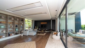Una sala de estar moderna con un sofá grande, una mesa de centro y una chimenea. Hay una estantería llena de objetos decorativos y un ventanal con vistas al exterior.