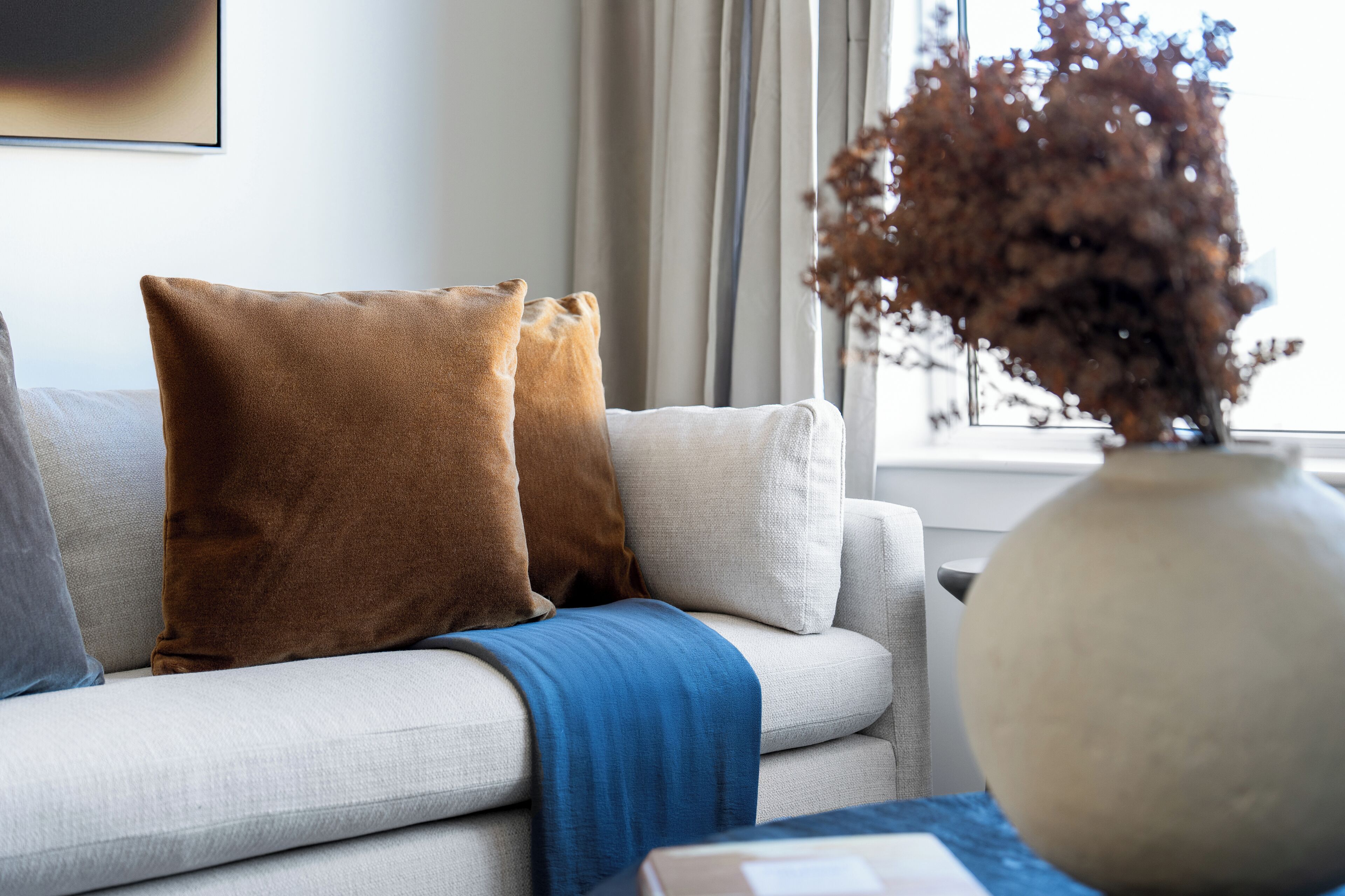 A sofa with two pillows, a blue throw blanket, and a vase with dried plants.