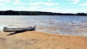Una canoa sobre una playa arenosa junto a un lago tranquilo con una orilla boscosa al fondo.