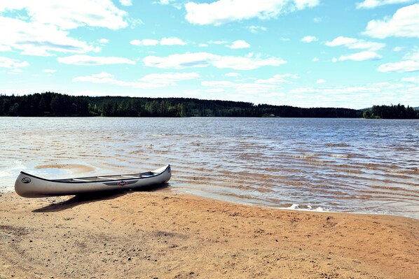 A canoe on a sandy beach by a calm lake with a forested shoreline in the background.