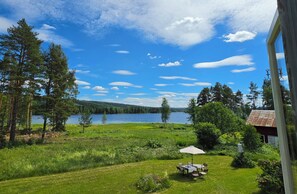 Näkymä järvelle, jossa on pöytä ja tuolit sijoitettu varjon alle, ja ympäristössä on vehreää kasvillisuutta sekä punakattoinen rakennus.