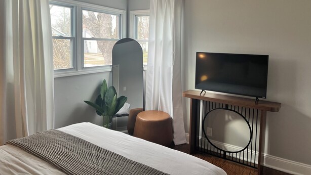 Une chambre d’hôtel moderne avec un lit, un téléviseur posé sur un meuble en bois, un grand miroir et une plante en pot.