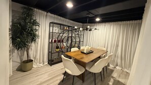 Un coin repas avec une table en bois, des chaises blanches, une plante en pot, une vitrine contenant des verres à vin et un lustre.