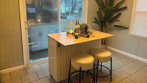 Un petit coin cuisine avec un plan de travail en bois, deux tabourets, une plante en pot et une fenêtre donnant sur une rue résidentielle.